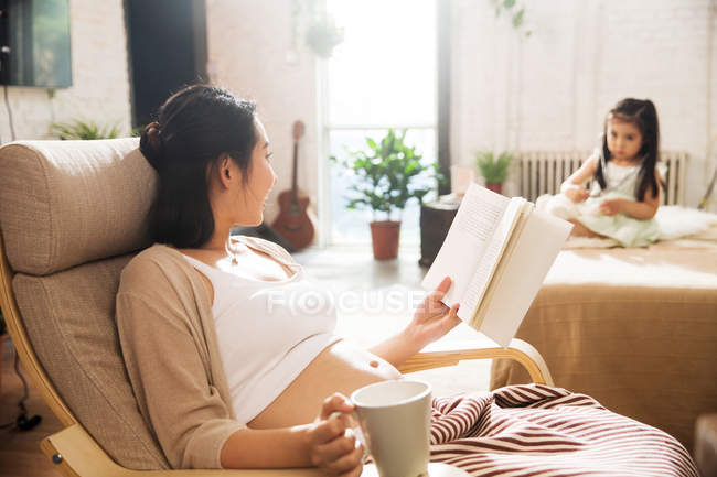 Jovem grávida segurando copo e leitura livro enquanto pequena filha brincando na cama — Fotografia de Stock