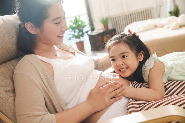 Очаровательный счастливый ребенок обнимает живот улыбающейся беременной матери дома — стоковое фото