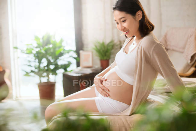 Вид сбоку улыбающейся молодой беременной женщины, сидящей на кровати и трогающей животик дома — стоковое фото