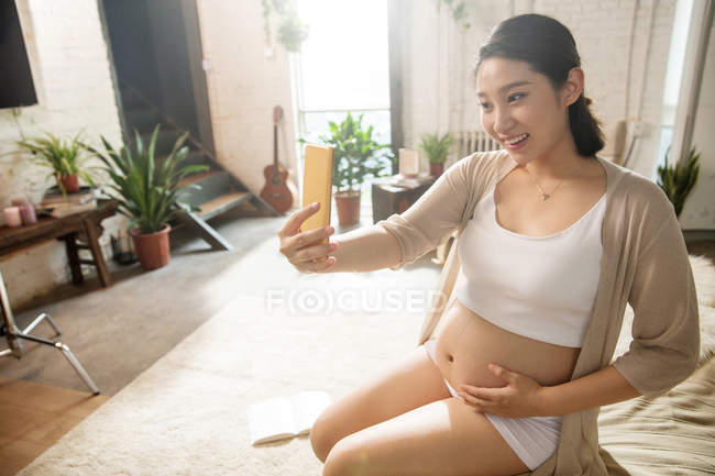 Blick aus der Vogelperspektive auf eine lächelnde junge Schwangere, die zu Hause ein Selfie mit dem Smartphone macht — Stockfoto