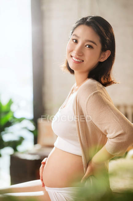 Lado vista de feliz joven embarazada asiático mujer sonriendo en cámara - foto de stock