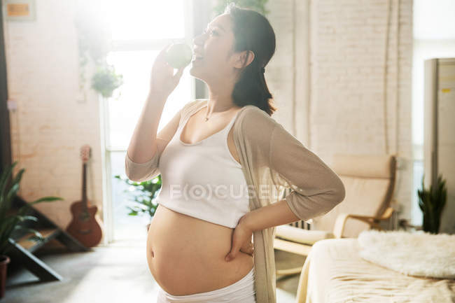 Vista lateral de feliz jovem grávida comendo maçã em casa — Fotografia de Stock