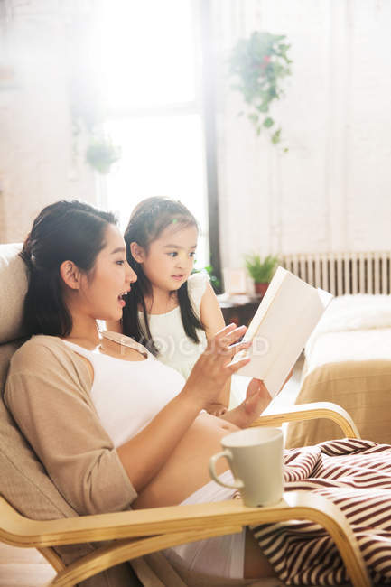 Embarazada joven china leyendo libro con adorable hijita en casa - foto de stock