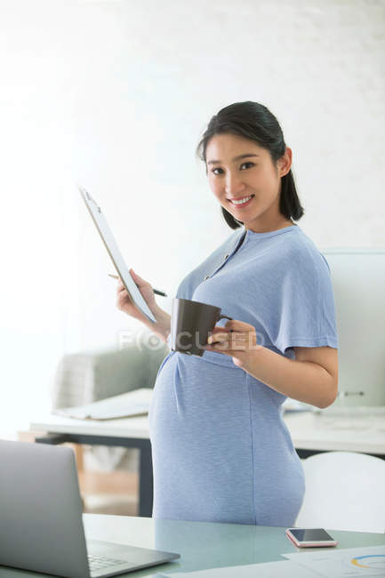 Jovem empresária grávida segurando prancheta e sorrindo para a câmera — Fotografia de Stock