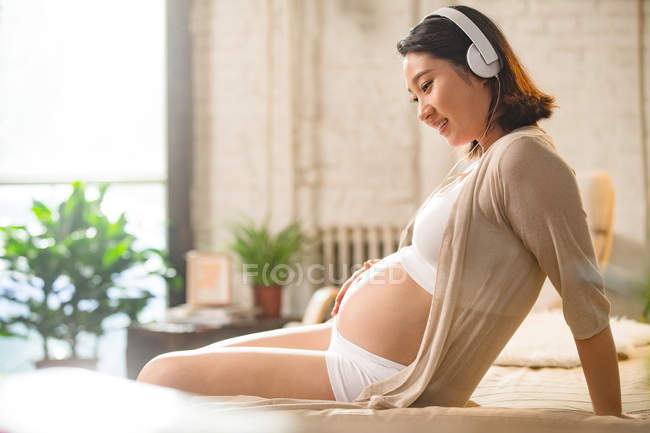 Visão lateral da mulher grávida sorrindo ouvir música em fones de ouvido em casa — Fotografia de Stock