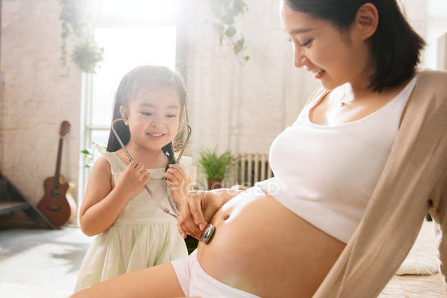 Entzückendes glückliches Kind hält Stethoskop in der Hand und hört auf den Bauch einer lächelnden Schwangeren — Stockfoto