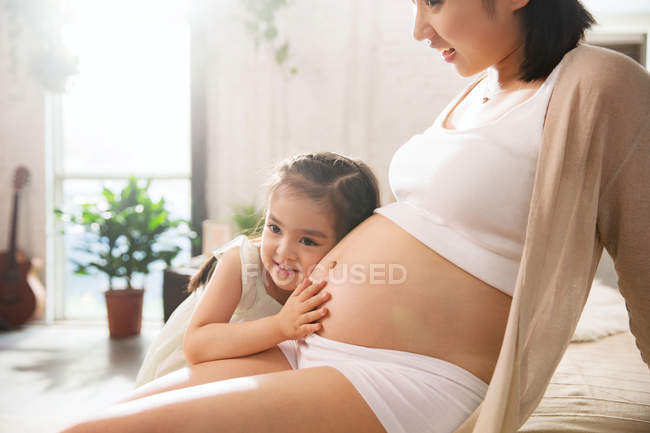Обрезанный снимок очаровательного счастливого ребенка, слушающего живот беременной матери — стоковое фото