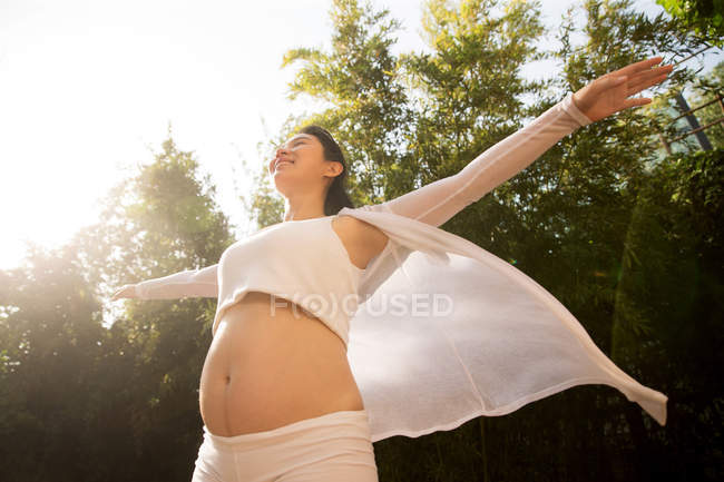 Vista de ángulo bajo de la joven embarazada feliz de pie con los brazos extendidos en el patio - foto de stock
