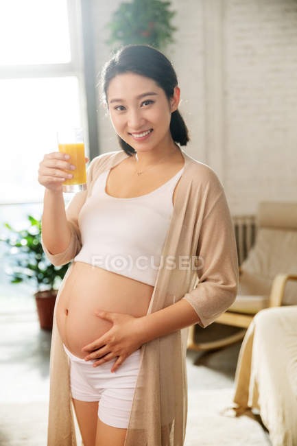 Feliz jovem grávida segurando vidro de suco e sorrindo para a câmera em casa — Fotografia de Stock