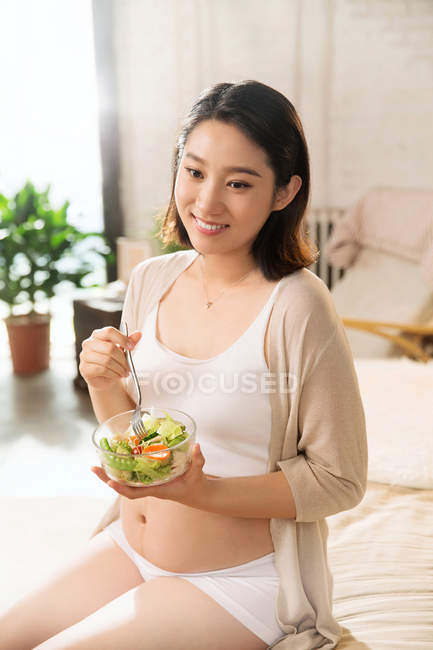 Улыбающаяся молодая беременная женщина сидит и держит миску с овощным салатом дома — стоковое фото