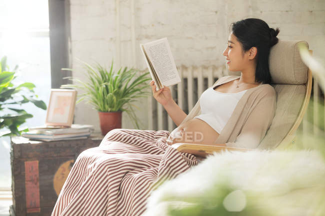 Vista lateral de la joven embarazada sonriente sentada en mecedora y leyendo libro en casa - foto de stock