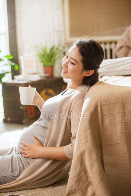Вид сбоку улыбающейся молодой беременной женщины, держащей дома чашку с горячим напитком — стоковое фото