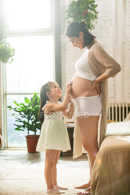Боковой вид очаровательного улыбающегося ребенка, держащего стетоскоп и слушающего живот беременной матери — стоковое фото