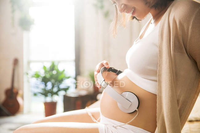 Обрезанный снимок улыбающейся беременной женщины, сидящей и держащей наушники на животе — стоковое фото