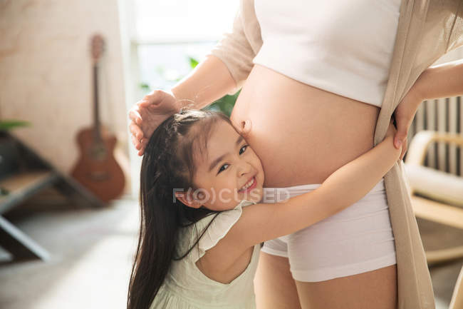 Обрізаний знімок чарівної щасливої дитини, що обіймає живіт вагітної мами і посміхається на камеру — стокове фото
