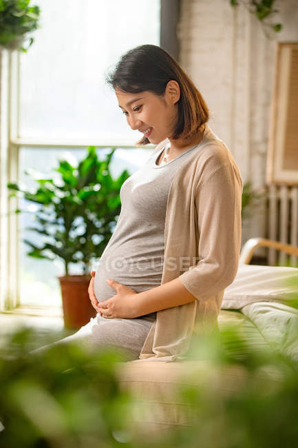 Side view of happy young pregnant asian woman sitting on bed and touching belly — Stock Photo