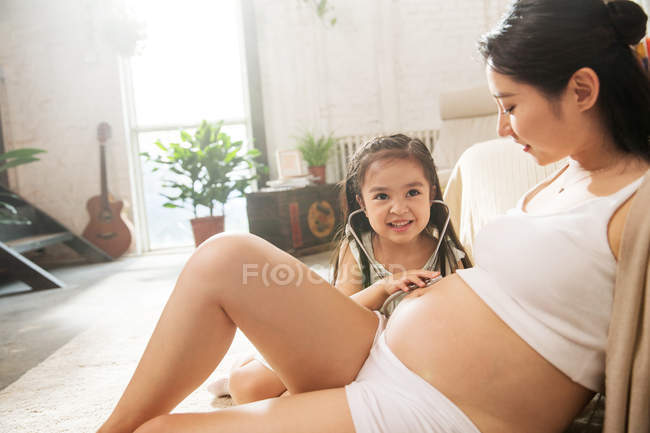 Adorable happy child holding stethoscope and listening belly of pregnant mother at home — Stock Photo