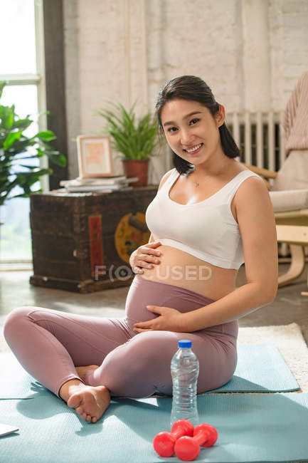 Feliz joven embarazada sentada en colchonetas de yoga, tocando el vientre y sonriendo a la cámara en casa - foto de stock