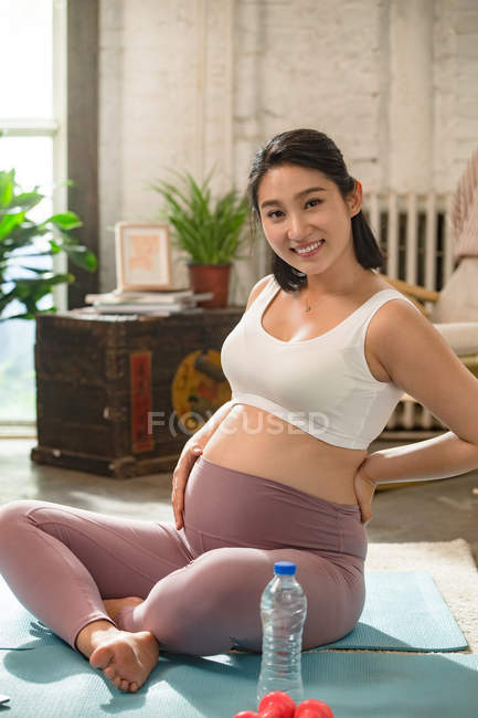 Щаслива молода вагітна жінка сидить на килимках йоги і посміхається на камеру вдома — стокове фото