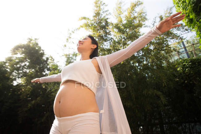 Низкий угол обзора счастливой молодой беременной женщины, стоящей с протянутыми во дворе руками — стоковое фото