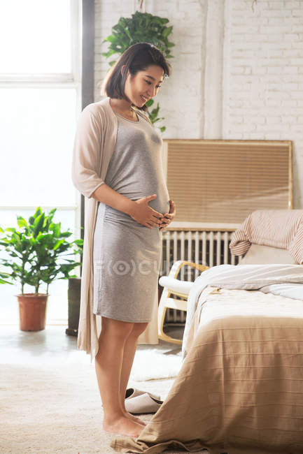 Feliz joven embarazada de pie en el dormitorio y tocando el vientre - foto de stock