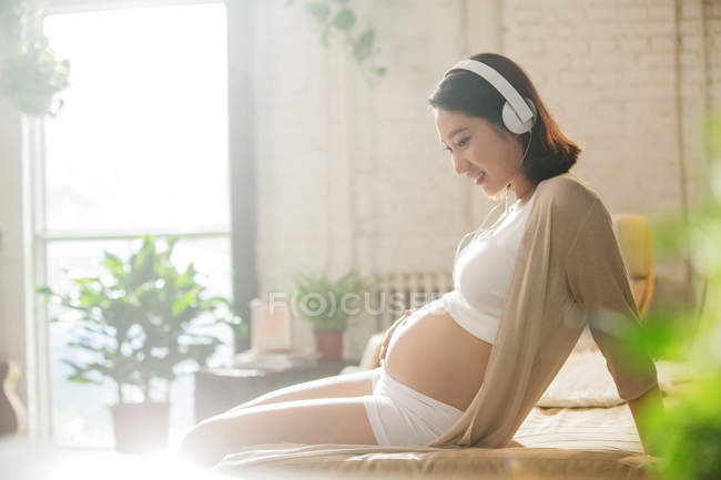 Side view of smiling young pregnant woman in headphones sitting on bed and touching belly — Stock Photo