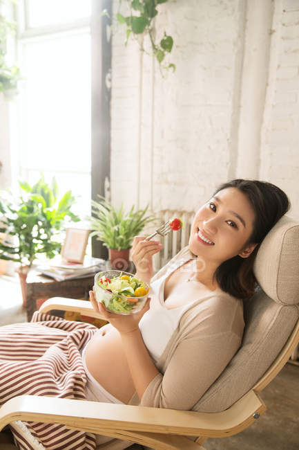 Молода вагітна жінка тримає миску з овочевим салатом і посміхається на камеру — стокове фото