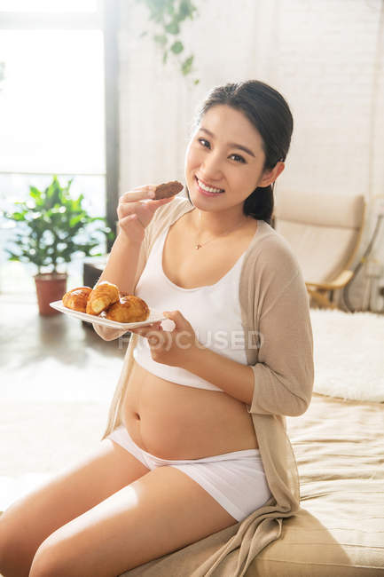 Happy young pregnant woman eating cookie and smiling at camera at home — Stock Photo