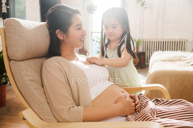Entzückendes Kind mit Stethoskop spielt mit schwangerer Mutter zu Hause — Stockfoto