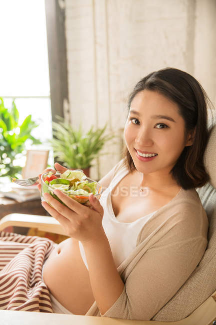Jeune femme enceinte tenant bol avec salade de légumes et souriant à la caméra — Photo de stock