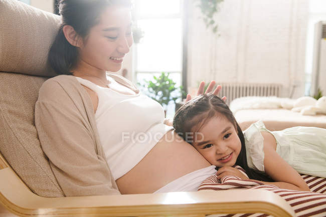 Очаровательный счастливый ребенок обнимает животик беременной матери дома — стоковое фото