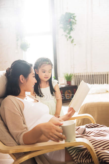 Pregnant young woman holding cup and reading book with adorable little daughter at home — Stock Photo