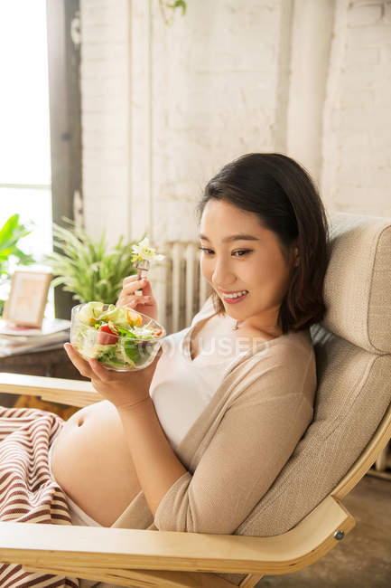 Счастливая молодая беременная женщина сидит в кресле и ест овощной салат дома — стоковое фото
