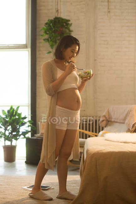 Lächelnde junge schwangere Asiatin steht und hält Schüssel mit Gemüsesalat zu Hause — Stockfoto