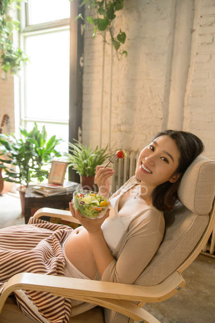 Giovane donna asiatica incinta mangiare insalata di verdure e sorridente a macchina fotografica a casa — Foto stock