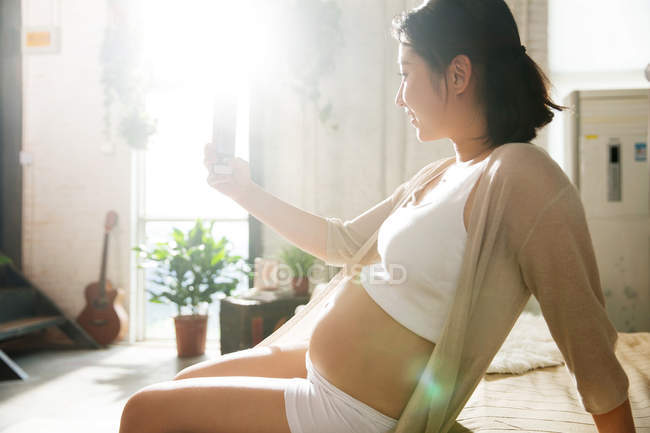 Боковой вид молодой беременной женщины, использующей смартфон дома — стоковое фото