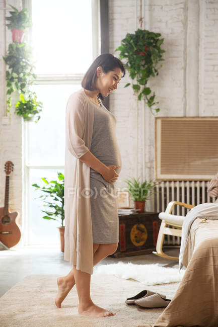 Side view of smiling young pregnant woman touching belly at home — Stock Photo