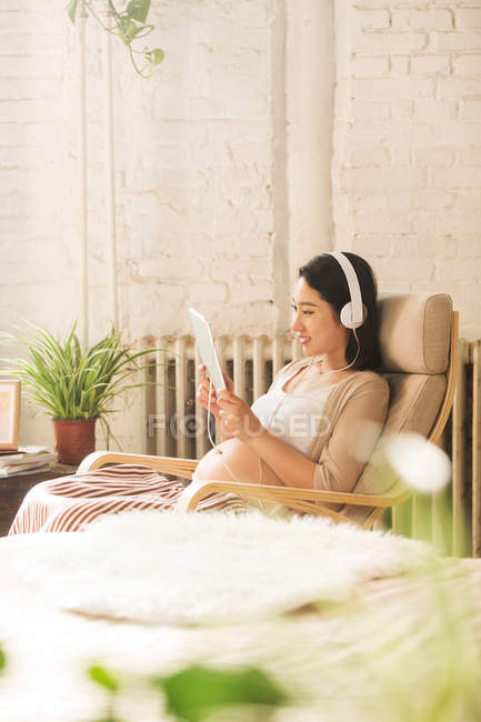 Souriant jeune femme enceinte dans les écouteurs en utilisant une tablette numérique à la maison — Photo de stock