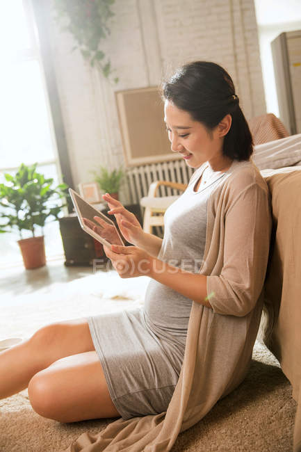 Боковой вид улыбающейся молодой беременной женщины, использующей дома цифровой планшет — стоковое фото
