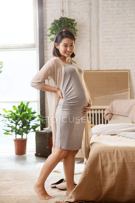 Hermosa feliz joven embarazada asiática mujer de pie y sonriendo en el dormitorio - foto de stock
