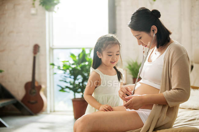 Чарівна щаслива дитина і вагітна мати грають разом зі стетоскопом вдома — стокове фото