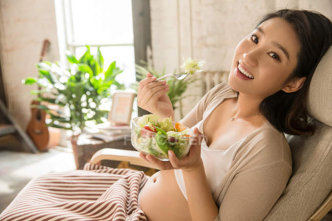 Glückliche junge schwangere Frau isst Gemüsesalat und lächelt zu Hause in die Kamera — Stockfoto
