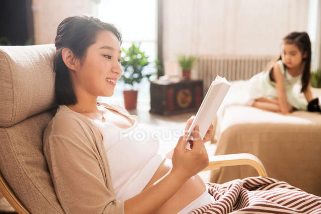 Sonriente joven embarazada leyendo libro mientras pequeña hija jugando en la cama - foto de stock