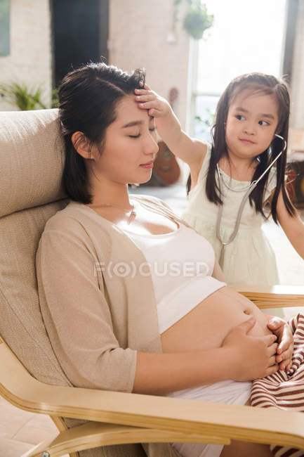 Entzückendes Kind spielt mit Stethoskop und gibt vor, Arzt zu sein, während schwangere Mutter im Stuhl sitzt — Stockfoto