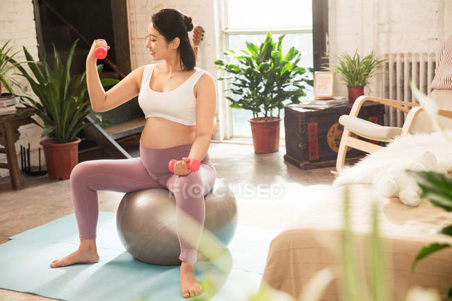 Young pregnant woman sitting on fit ball and exercising with dumbbells at home — Stock Photo