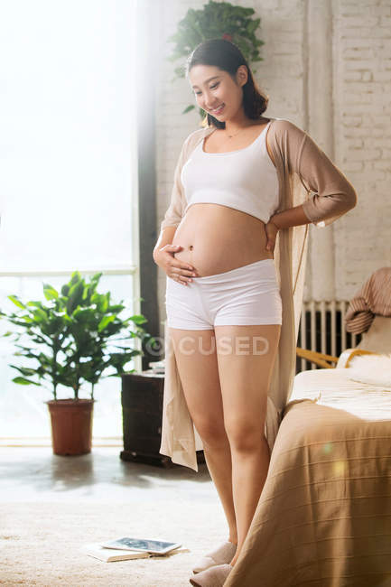 Щаслива молода вагітна жінка стоїть і посміхається в спальні — стокове фото