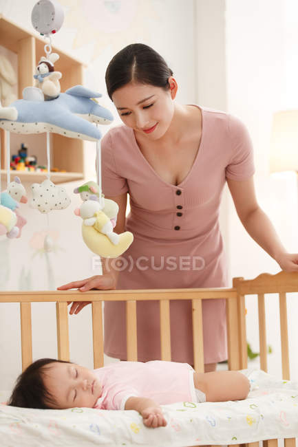 Улыбающаяся молодая китайская женщина смотрит на очаровательного ребенка, спящего в кроватке — стоковое фото