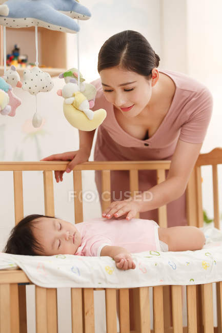Улыбающаяся молодая китайская женщина смотрит на очаровательного ребенка, спящего в кроватке — стоковое фото
