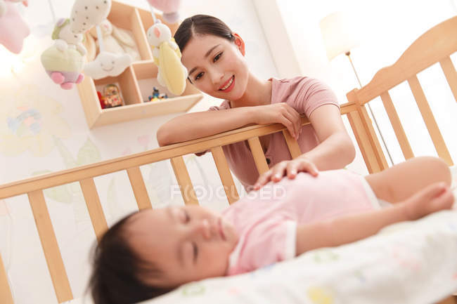 Glückliche junge Mutter blickt auf entzückendes Baby, das im Kinderbett schläft — Stockfoto