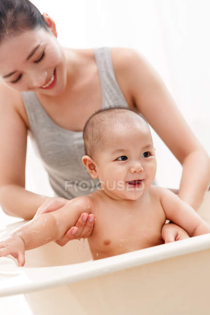 Happy young mother bathing adorable smiling infant baby sitting in bathtub — Stock Photo